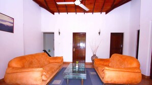 A living room with two orange sofas, a glass coffee table, and a ceiling fan. - The Blue Beach Guesthouse (Negombo)
