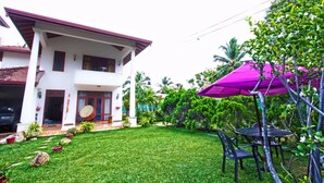 A two-story house with a white exterior and a red roof. There is a patio with a purple umbrella and a table with chairs. - The Blue Beach Guesthouse (Negombo)