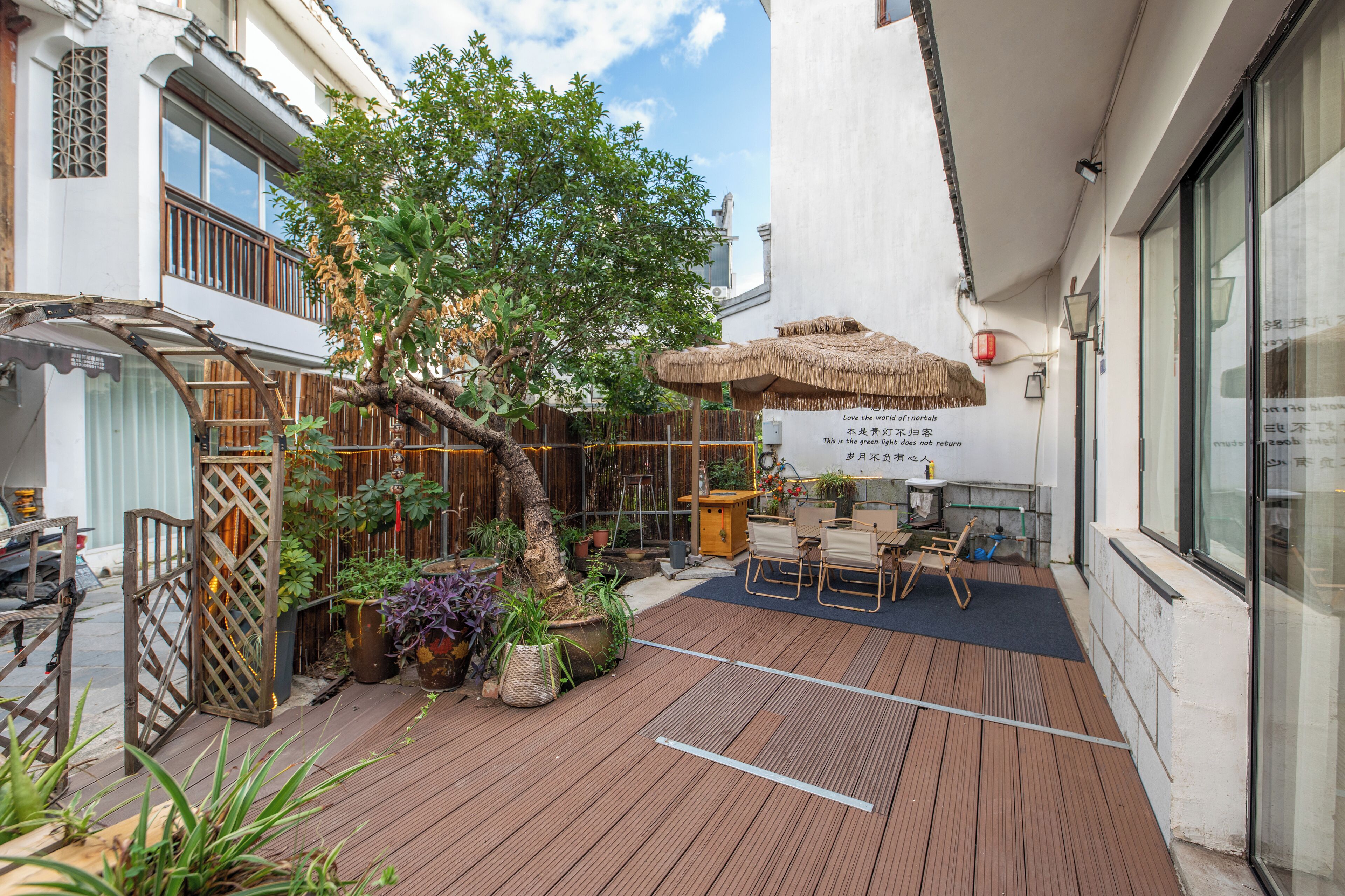 Un área de patio al aire libre con terraza de madera, un paraguas de paja y diversas plantas en macetas.