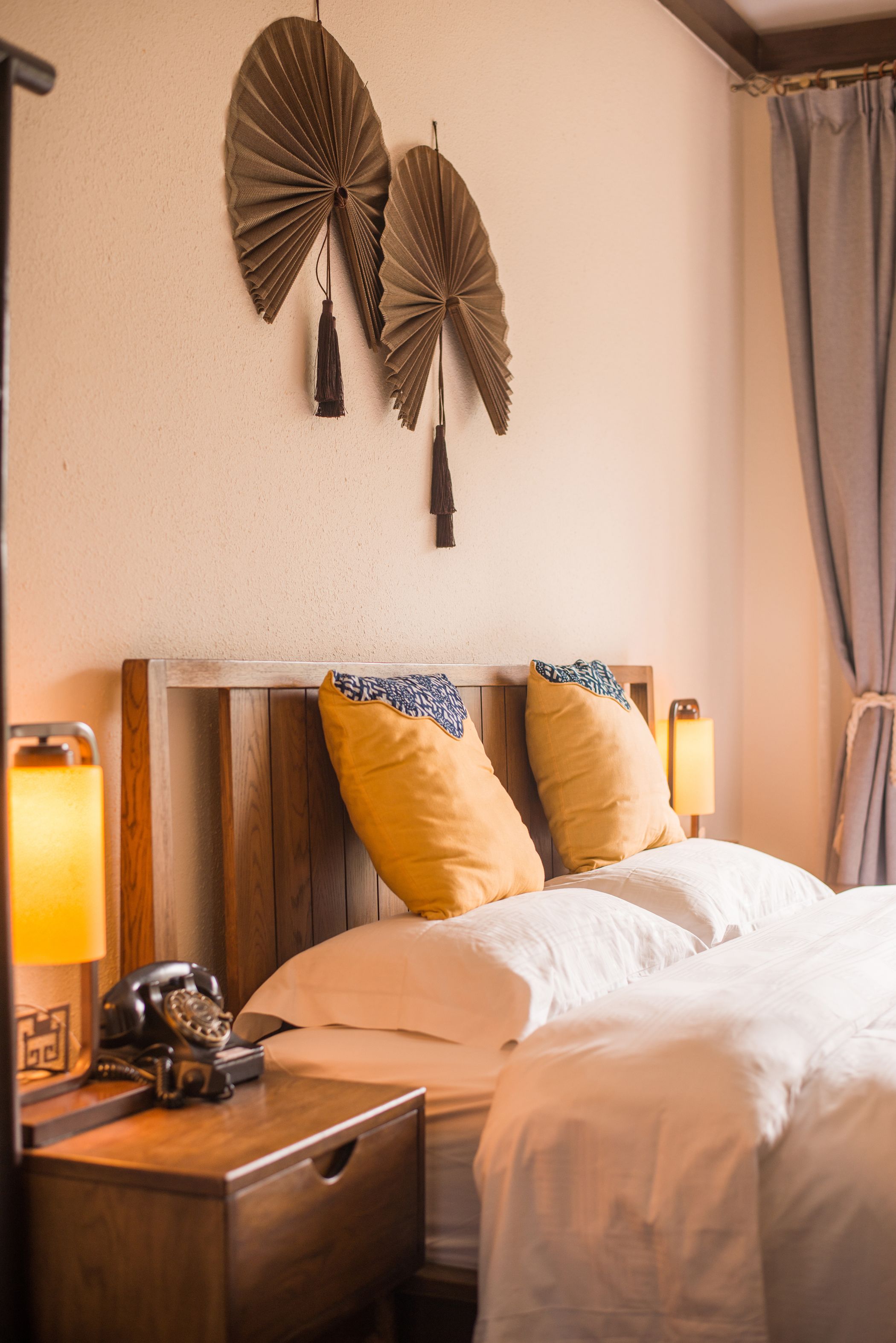 A hotel room with a wooden headboard, two yellow pillows, a white bedspread, a bedside lamp, and a vintage telephone on the nightstand.