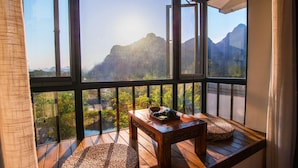 A wooden deck with a table and woven poufs, overlooking a mountainous landscape through large windows. - Secret Courtyard Resort Hotel (Guilin)