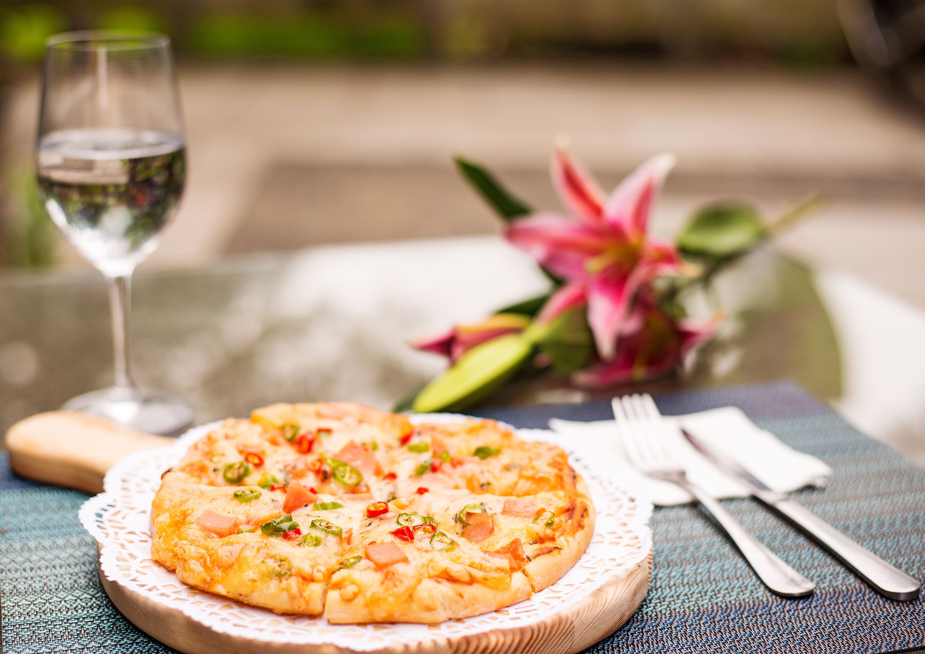 A pizza on a wooden board with a lace doily, a glass of water, a fork, a knife, and a pink flower in the background.