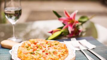 A pizza on a wooden board with a lace doily, a glass of water, a fork, a knife, and a pink flower in the background.