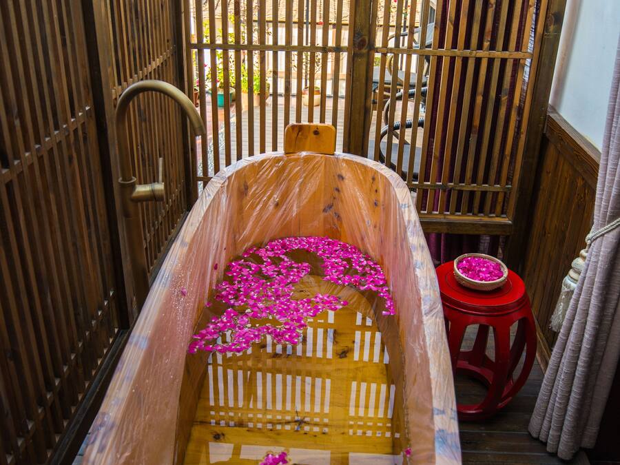 Una bañera de madera llena de agua y flores rosas, un taburete rojo y un portón de madera al fondo.