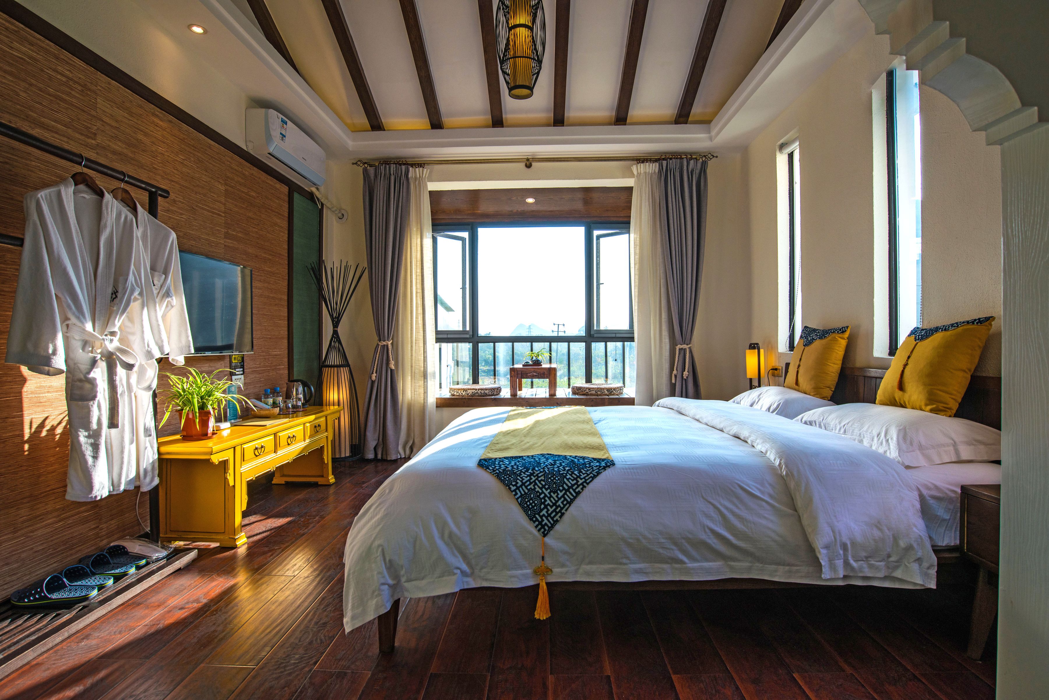 A spacious bedroom with a large bed, a wooden dresser, a vanity with a mirror, and a window with a view.