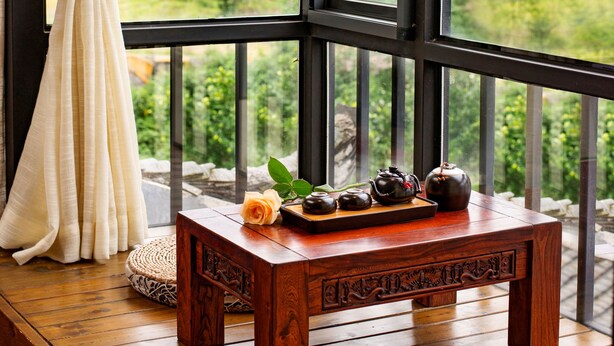 A wooden table with a tea set and a vase of flowers on a balcony with a view of green hills and a mountain. - Secret Courtyard Resort Hotel (Guilin)
