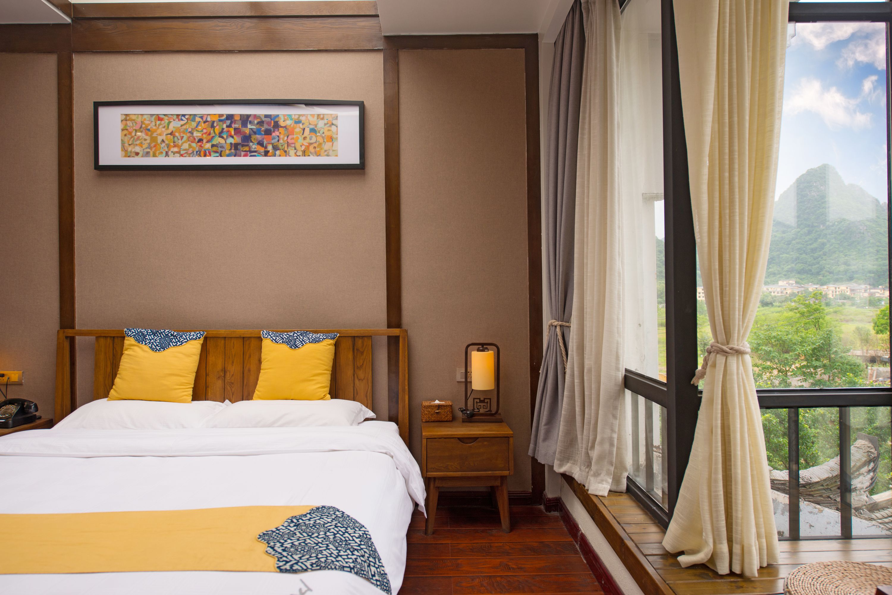 A hotel room with a bed, a bedside table with a lamp, a framed artwork on the wall, and a window with curtains.