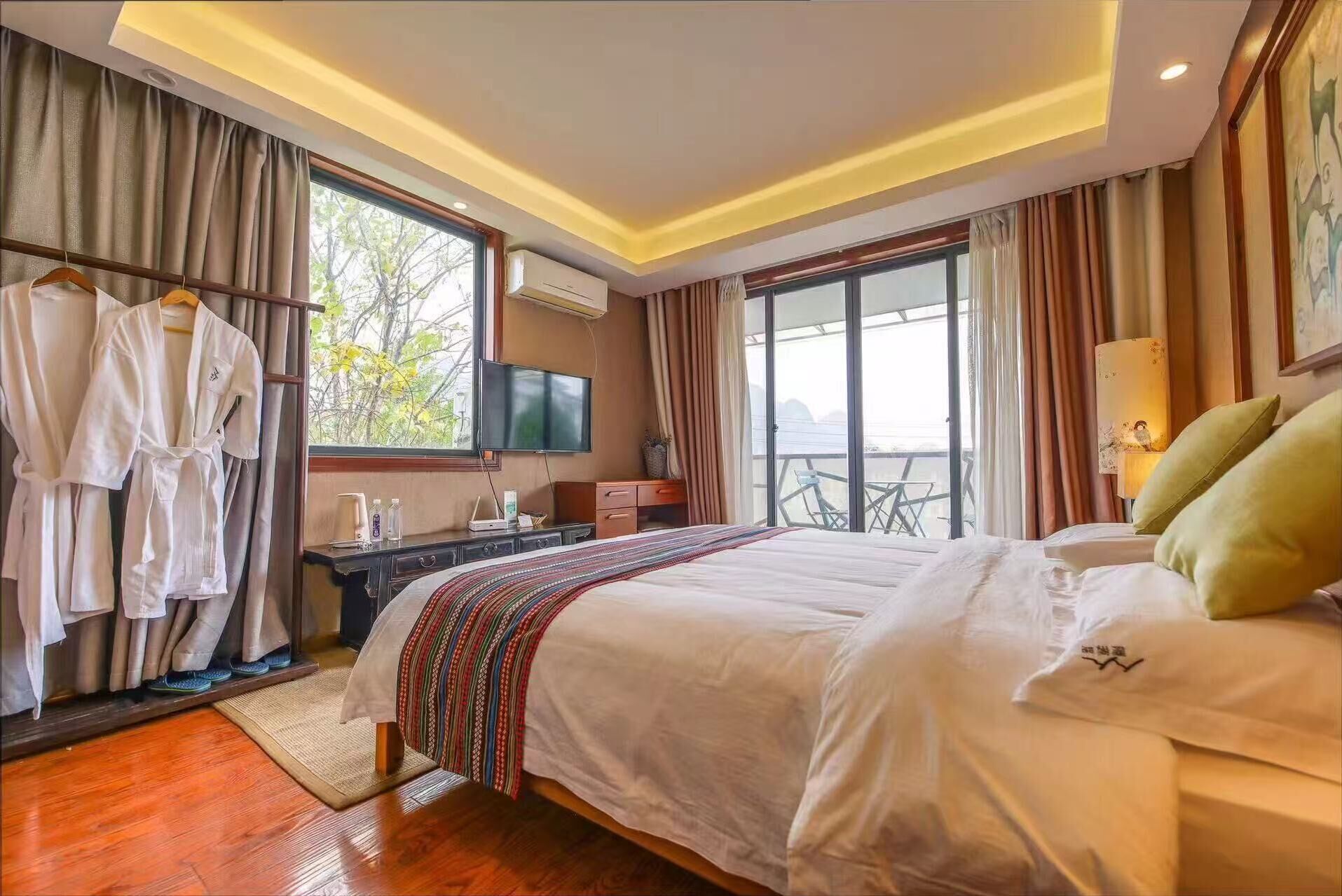 A hotel room with a large bed, a desk with a flat-screen TV, a balcony with a table and chairs, and a robe rack with white robes.