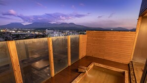 A wooden deck with a hot spring bath and a cityscape view in the background. - Aso Hotel (Aso)