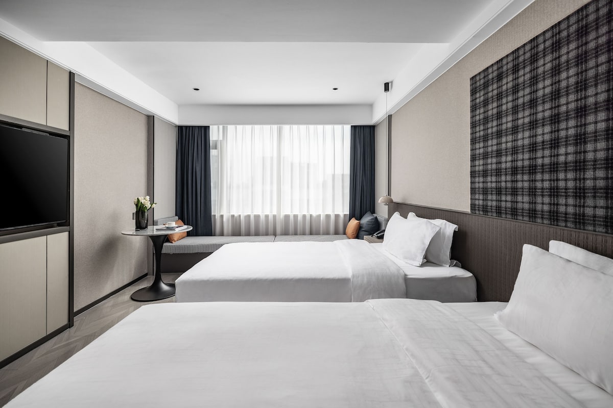 A modern hotel room with a large bed, a flat-screen TV, a small table with a vase, and a large window with curtains.