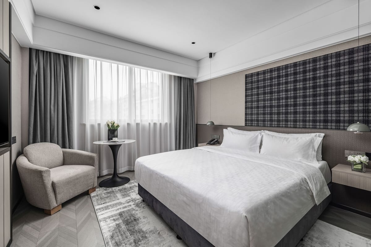 A modern hotel room with a large bed, a seating area with a chair, a small table, and a bedside lamp.