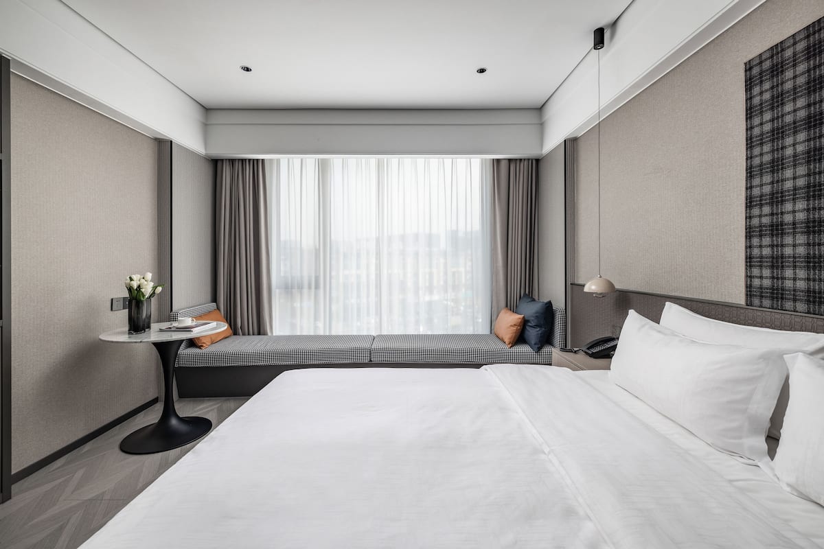 A hotel room with a large bed, a small table with a vase of flowers, a sofa with cushions, a telephone, and a window with curtains.