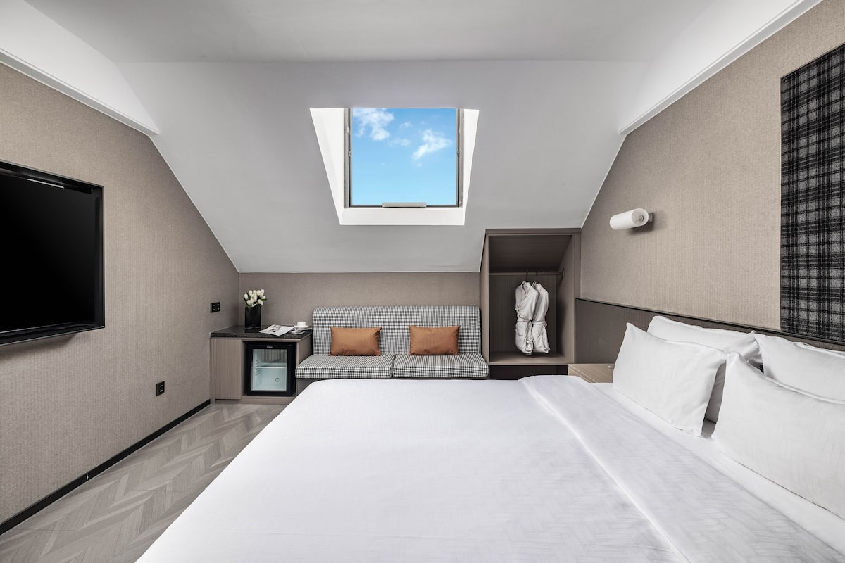 A modern hotel room with a large bed, a TV, a small desk, a sofa, and a wardrobe.