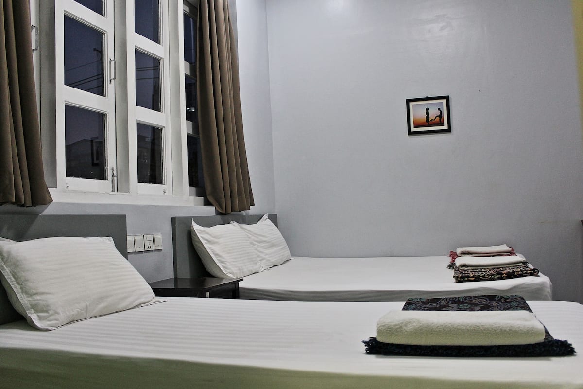 Habitación de hotel con dos camas individuales, una ventana con cortinas, un pequeño cuadro en la pared y una cama bien hecha con una toalla doblada.