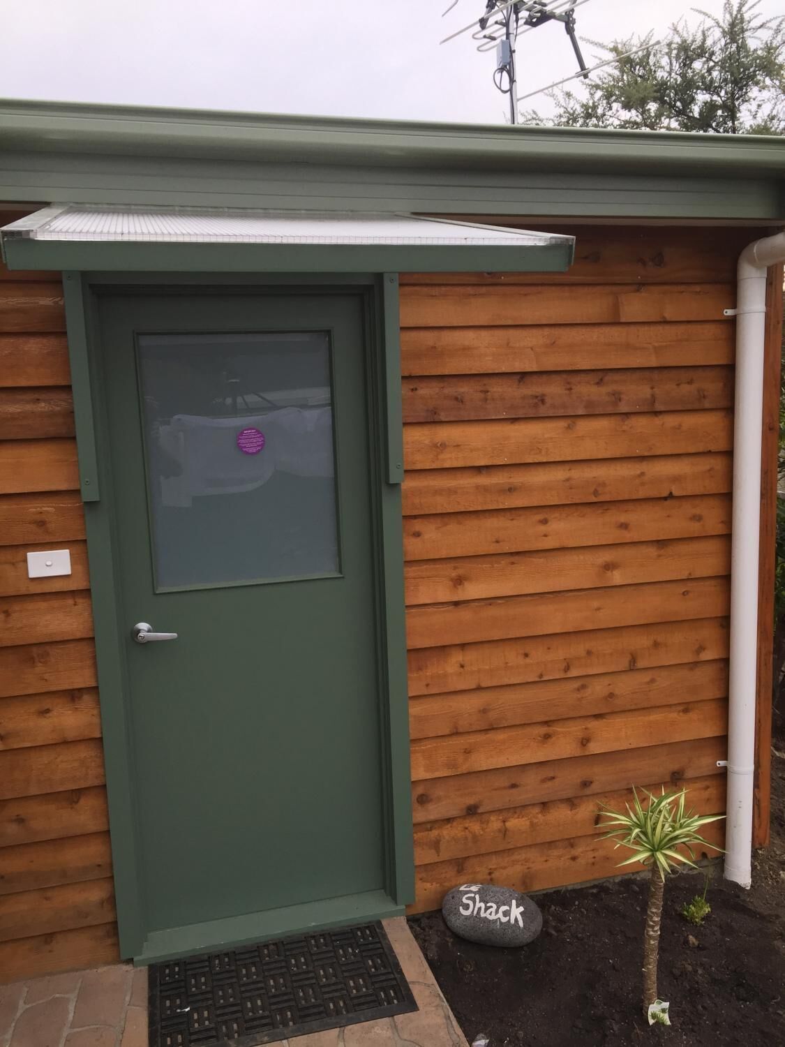 A green door with a window, a small stone labeled "Shack," and a palm tree in front of a wooden wall.