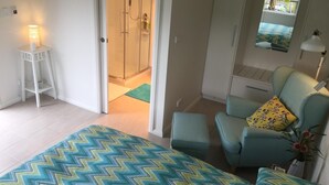 Un dormitorio con una cama, una silla, una mesita y un baño visible a través de una puerta abierta.