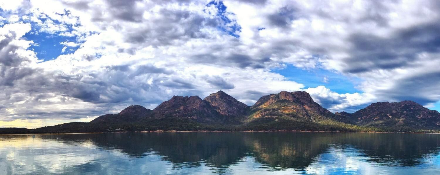 A mountain range reflected in calm water under a partly cloudy sky.