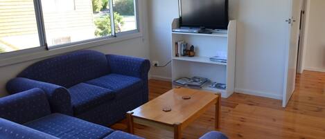 Uma sala de estar com um sofá azul, uma mesa de centro de madeira e uma televisão sobre um suporte branco.