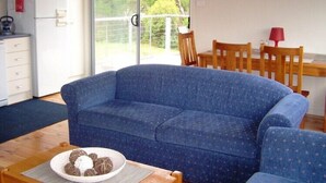 Sala de estar com sofá azul, mesa de jantar com cadeiras, cozinha com frigorífico e micro-ondas, e uma grande porta de vidro deslizante que dá para uma varanda.