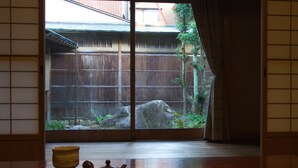 Ein traditioneller japanischer Raum mit Tatami-Boden, Shoji-Schirm, einem Tee-Set und Blick auf einen Garten mit Holzzaun und Baum.
