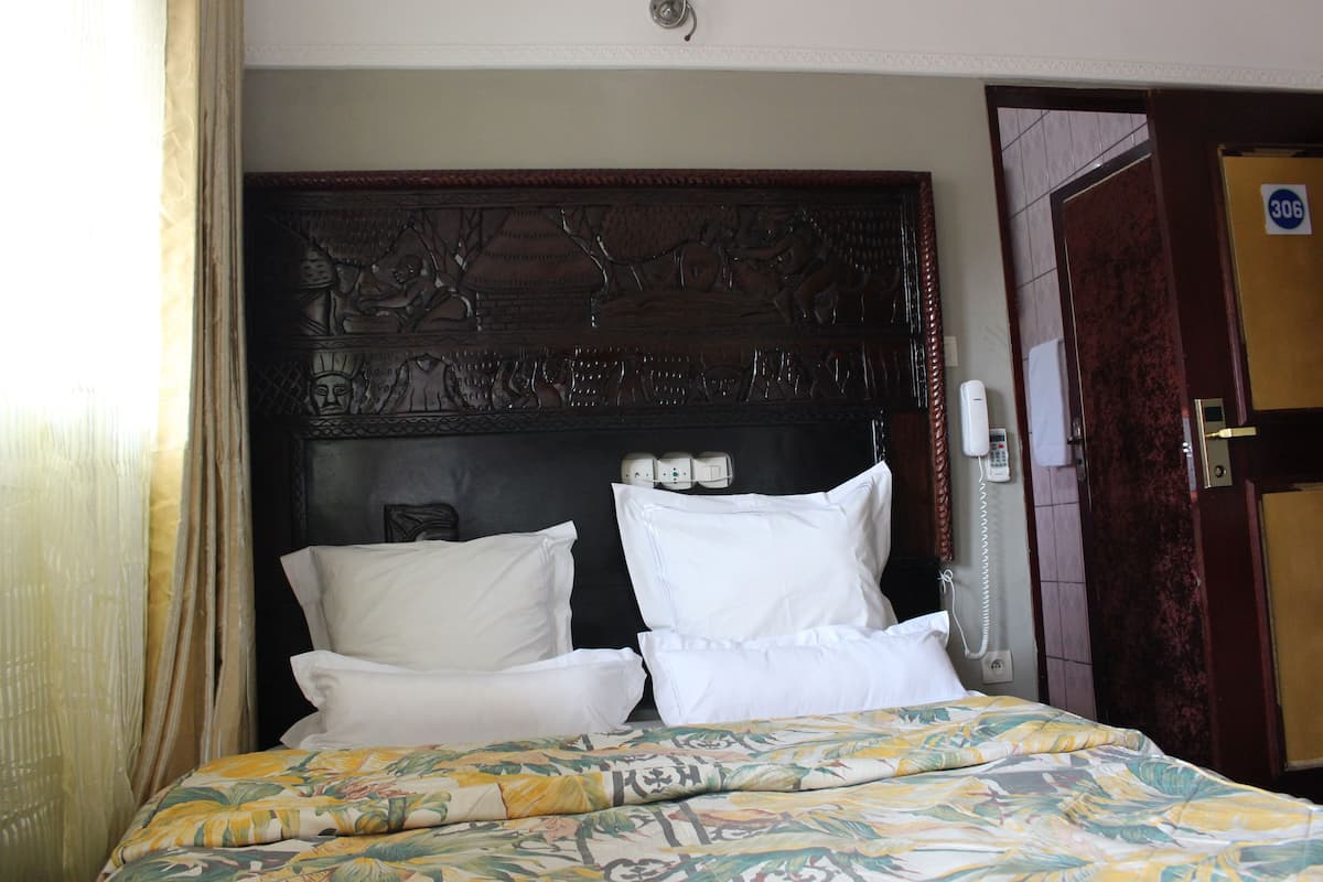 A hotel room with a bed, white pillows, a floral bedspread, a dark headboard with carvings, a telephone, and a door with a number sign.