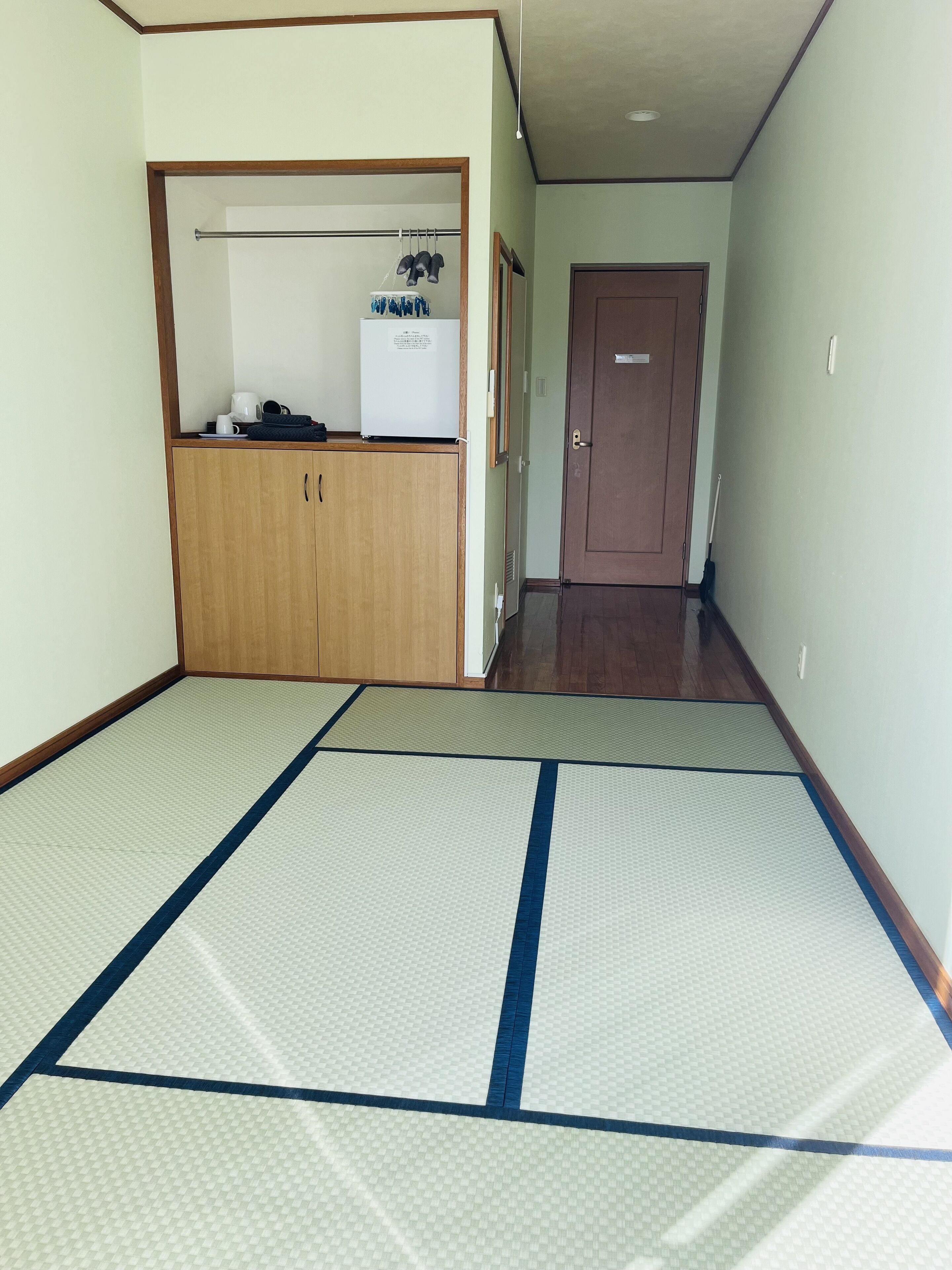A room with tatami mats, a wooden wardrobe, a small refrigerator, and a door in the background.