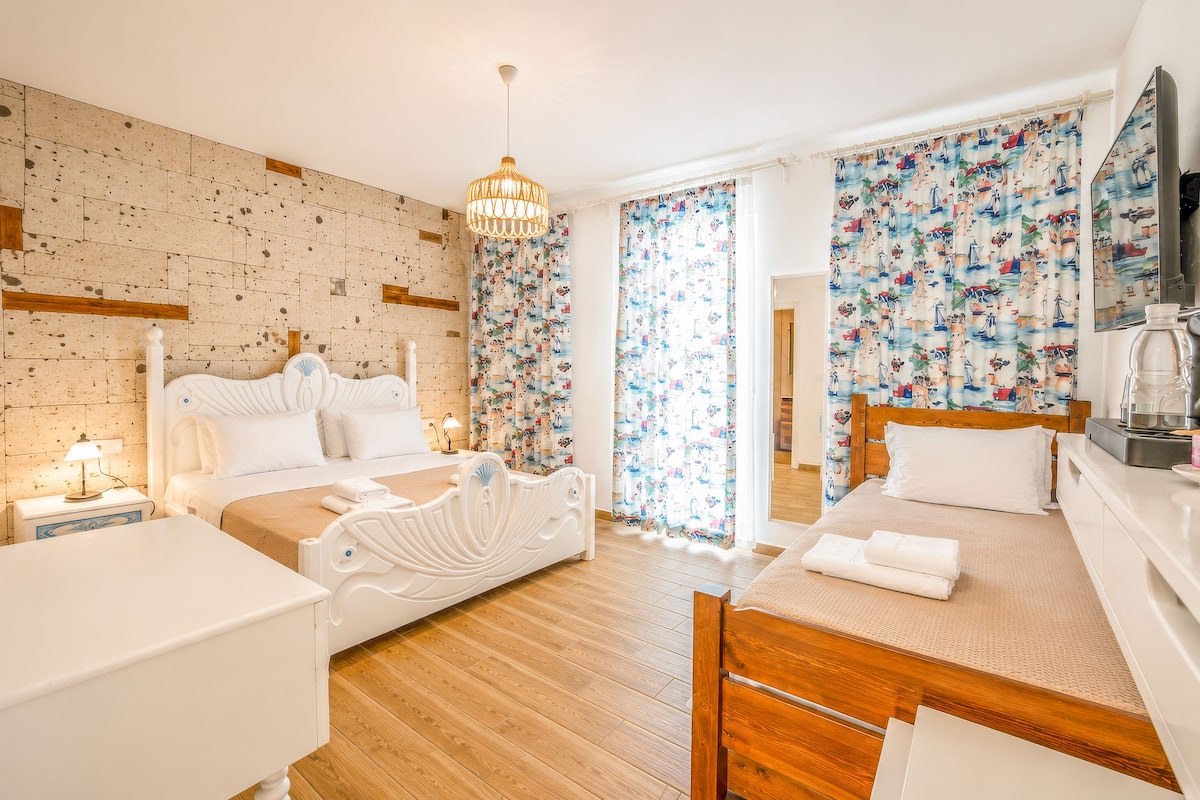 A hotel room with a wooden floor, a white bed with a headboard, a bedside table with a lamp, and a large window with curtains.