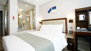 Ein Hotelzimmer mit einem großen Bett, einer Duschkabine aus Glas, einem Nachttisch aus Holz mit Lampe sowie einer Minibar.