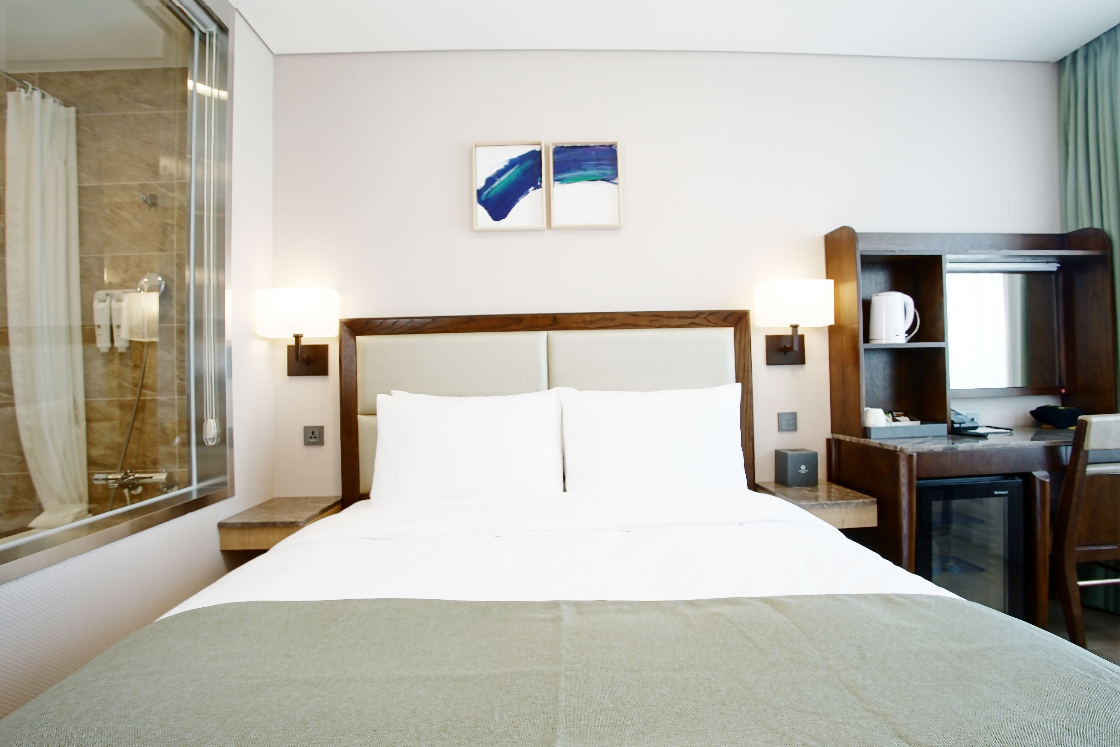 Habitación de hotel con una cama grande, un escritorio, una silla, un refrigerador pequeño, un espejo y zona de ducha.