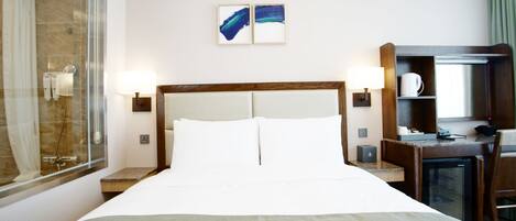 Habitación de hotel con una cama grande, un escritorio, una silla, un refrigerador pequeño, un espejo y zona de ducha.