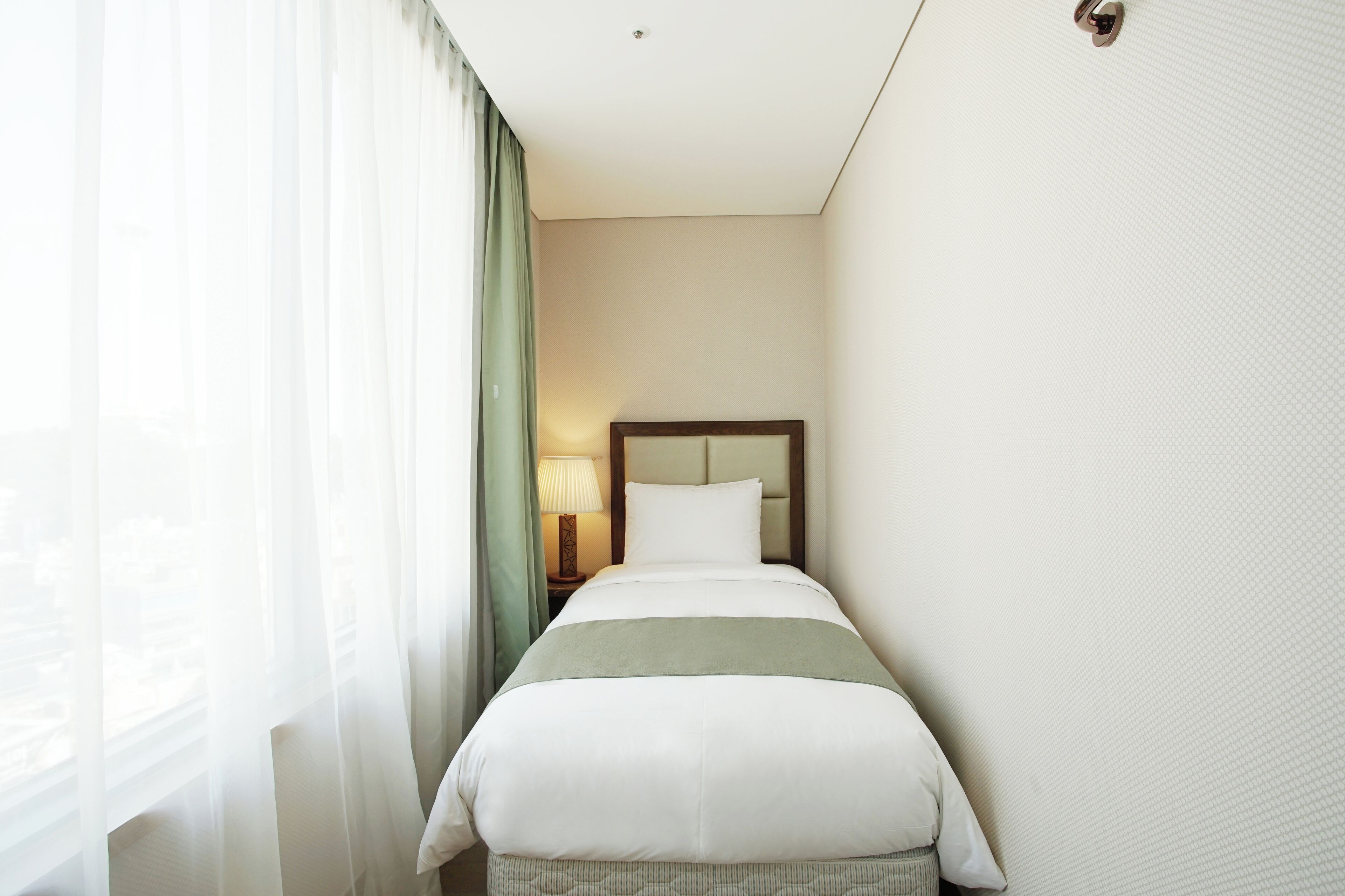 A hotel room with a bed, a nightstand with a lamp, and a window with sheer curtains.