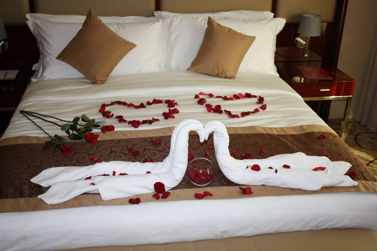 Un lit bien fait, avec une literie blanche et marron, un verre d’eau et des pétales de rose disposés sur le lit.
