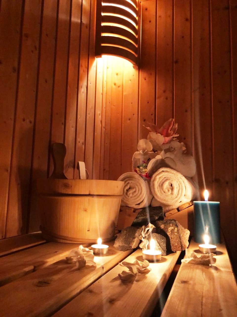 A wooden sauna with a bench, a wooden bucket, rolled towels, and lit candles.