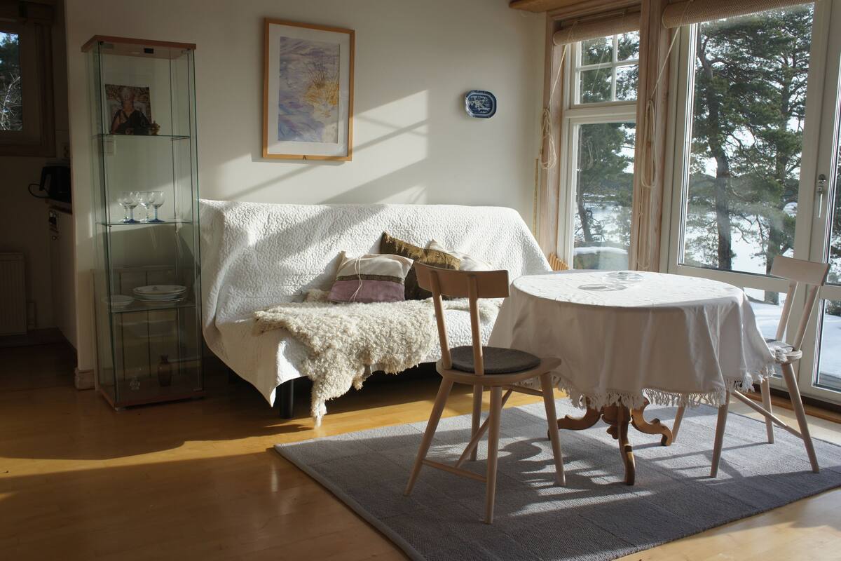 Ein gemütliches Zimmer mit einem weißen Sofa, einer Glastruhe, einem runden Tisch und Blick auf Bäume durch ein Fenster.