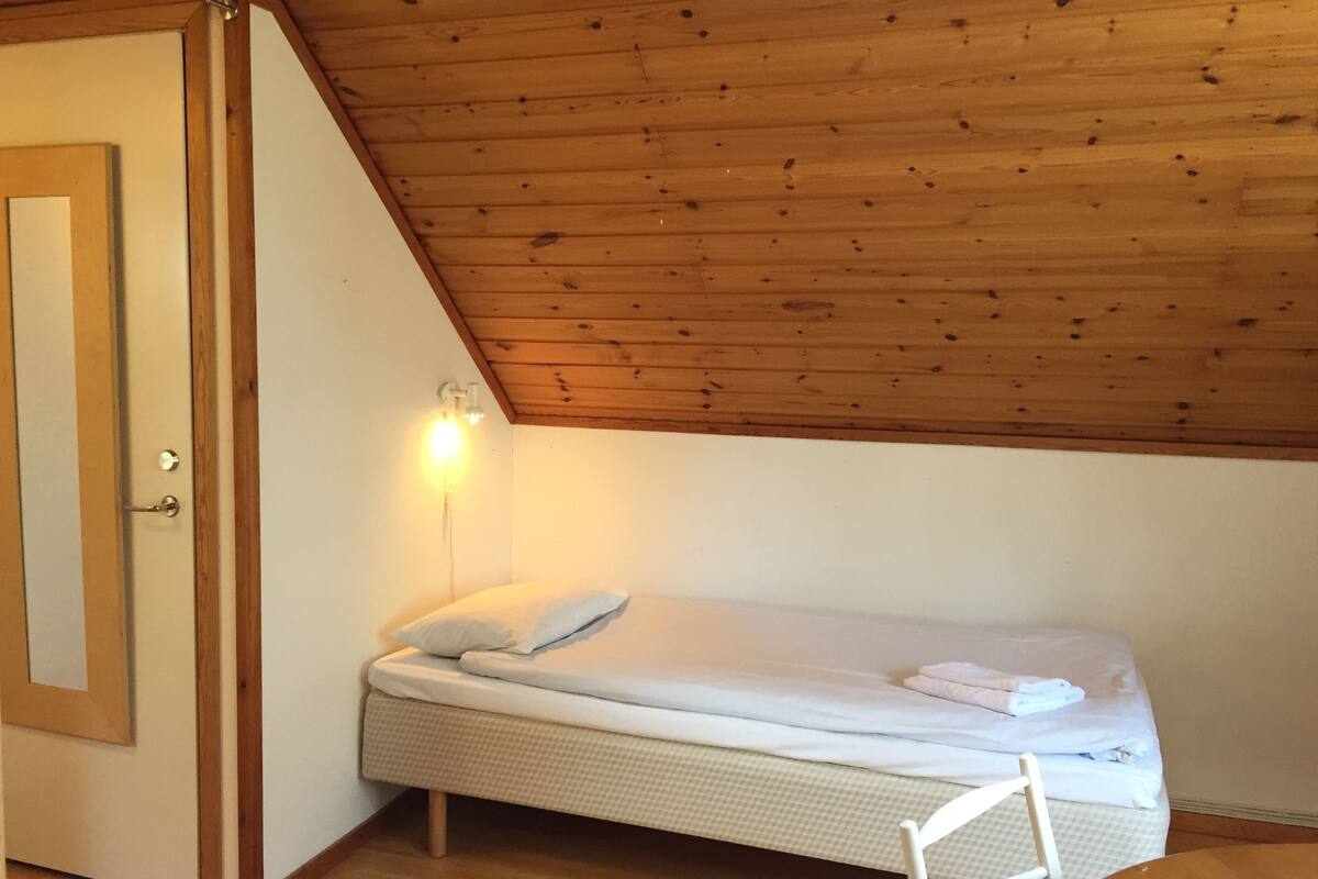 Ein kleines Zimmer mit einem Einzelbett, geneigter Decke und Holzbalken. Es gibt einen Nachttisch mit Lampe, eine Tür mit Spiegel und einen Stuhl.