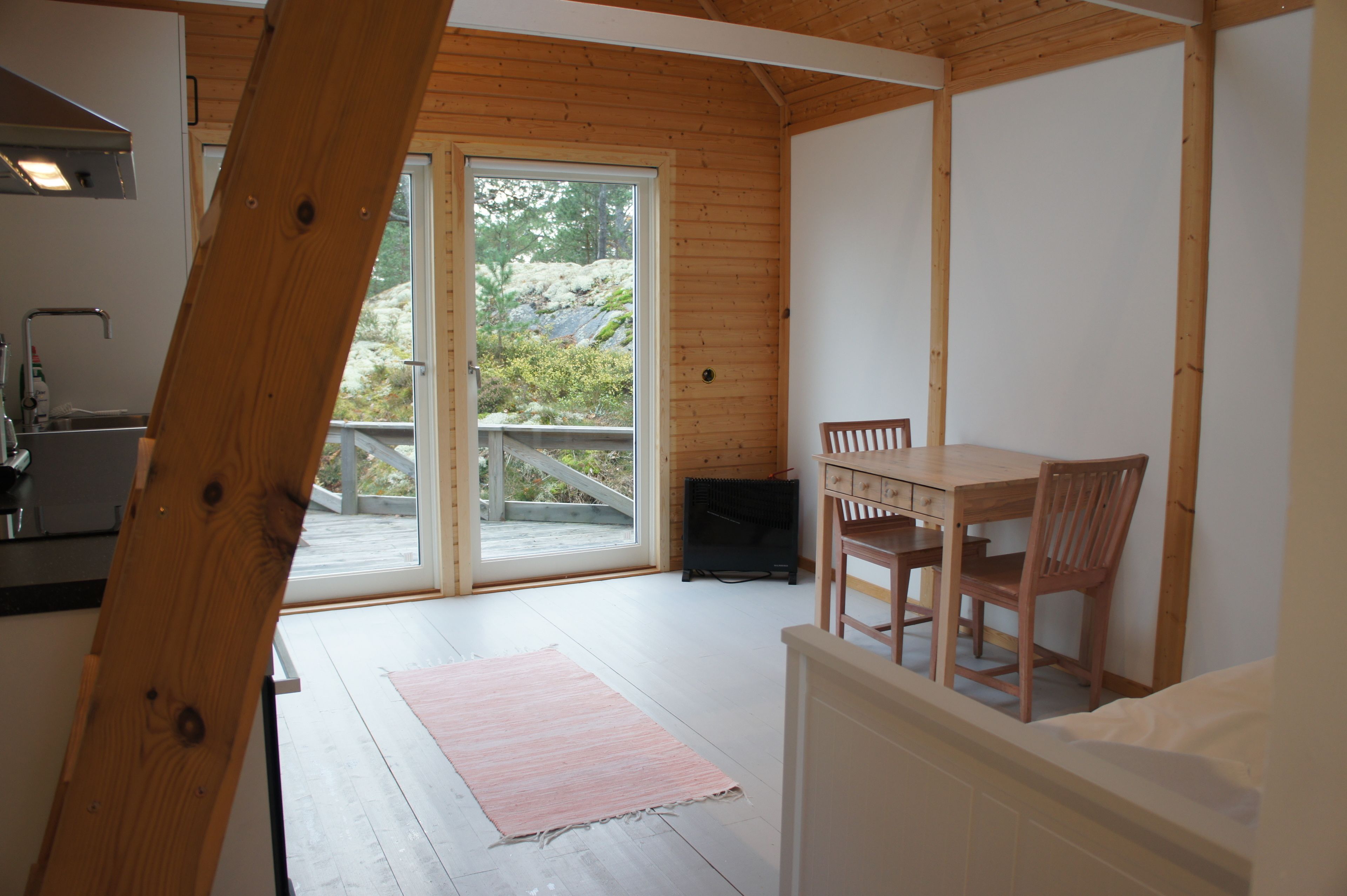 Una habitación con techo de madera, una mesa de comedor con sillas, un tapete rosa, una cama y un ventanal corredero que da a un espacio exterior con vistas a árboles y rocas.