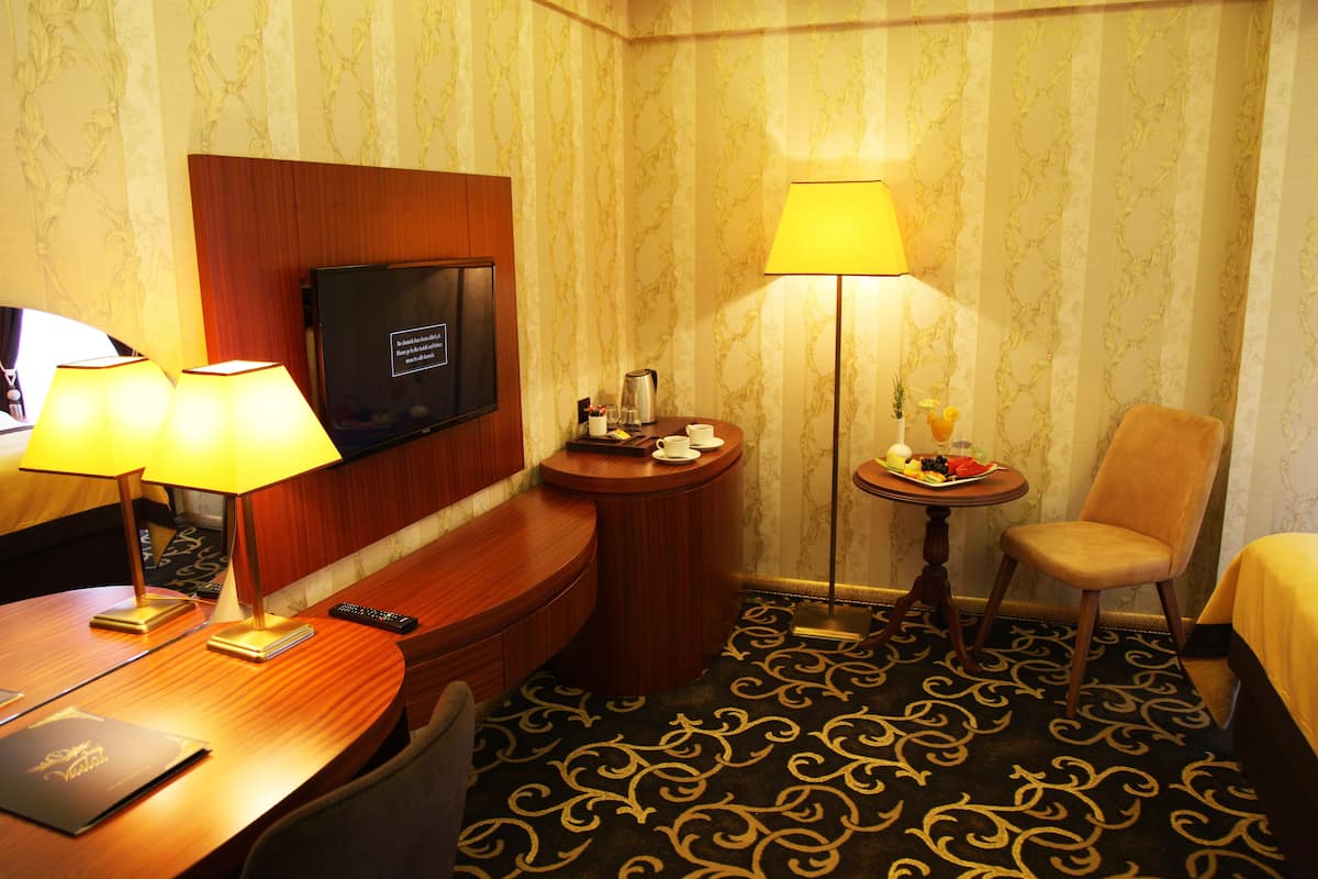 A hotel room with a bed, a desk with a lamp, a television, a small table with a fruit plate, and a chair.
