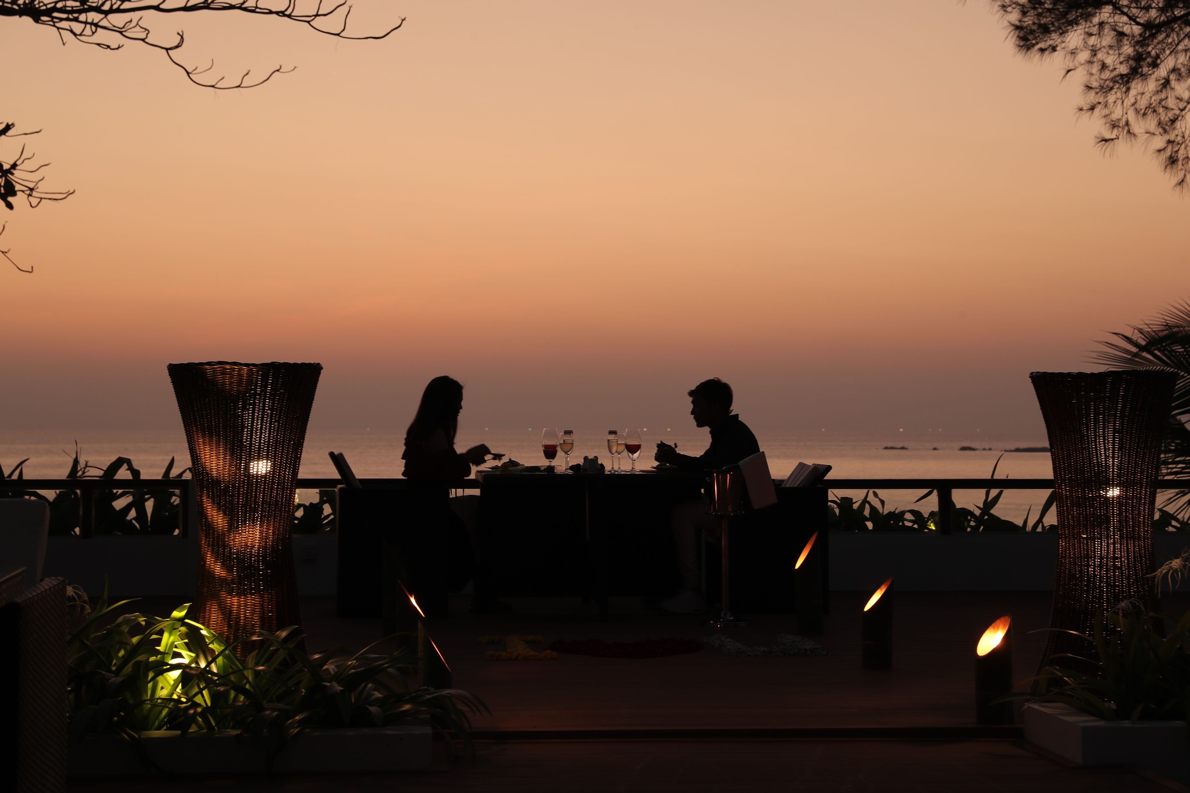 Une vue sur le coucher de soleil depuis un espace de restauration en plein air, avec des tables et des chaises, entouré de plantes et d’une rambarde.