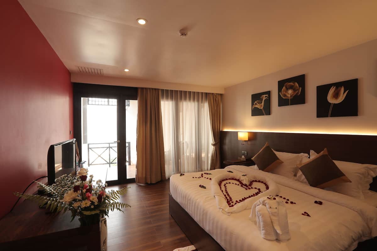 Quarto de hotel com uma cama, uma escrivaninha com um vaso de flores, uma televisão e uma porta de correr para uma varanda.