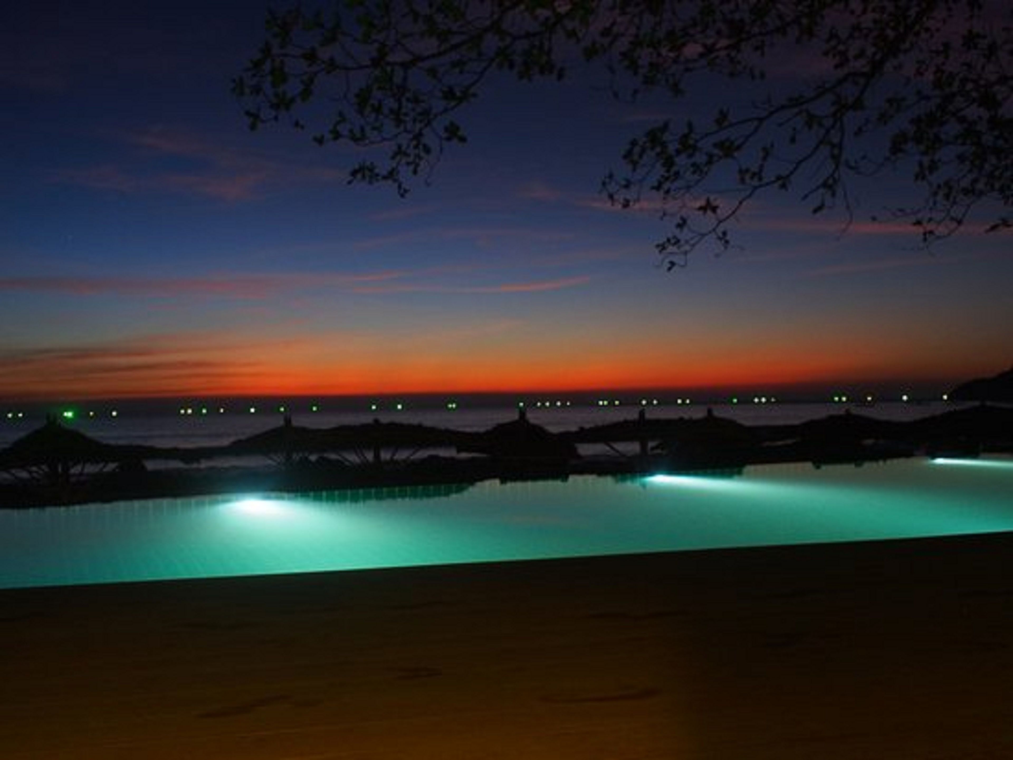 A nighttime view of a waterfront with a pool reflecting lights, a sunset in the background, and distant city lights.