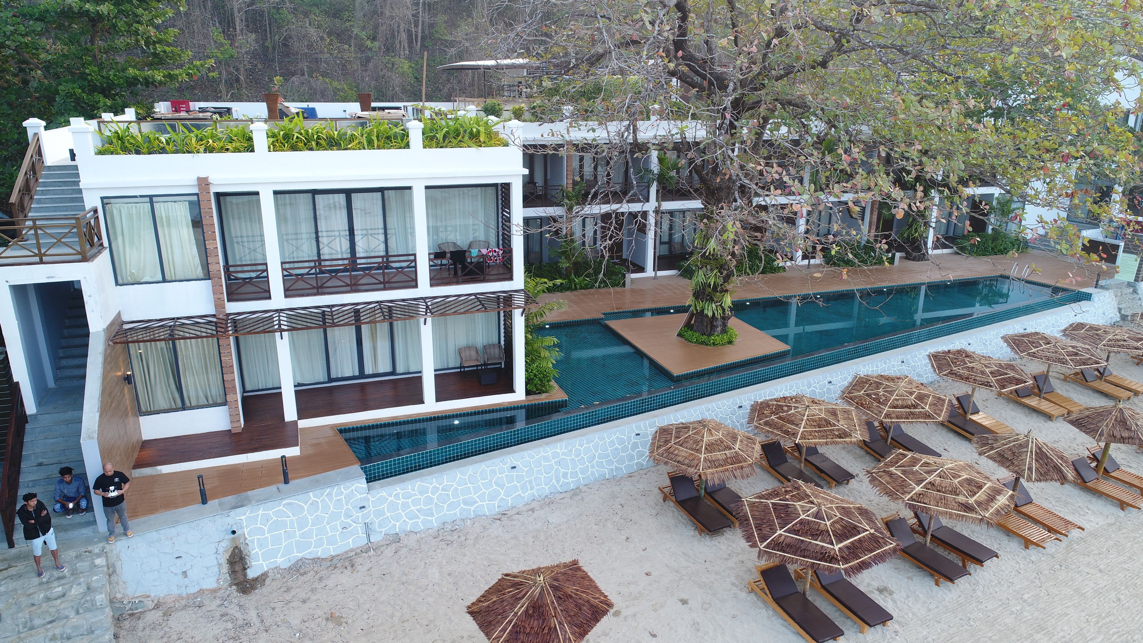 A modern hotel with a rooftop pool, multiple balconies, and a beach area with sun loungers.