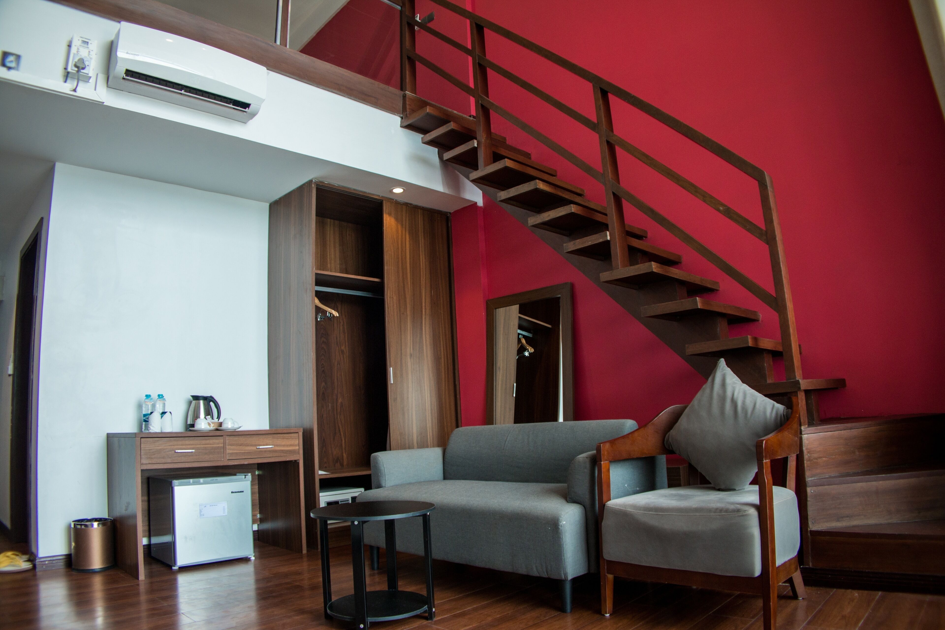 Una sala de estar moderna con un sofá gris, una silla de madera, una mesita, un armario empotrado y una escalera frente a una pared roja.