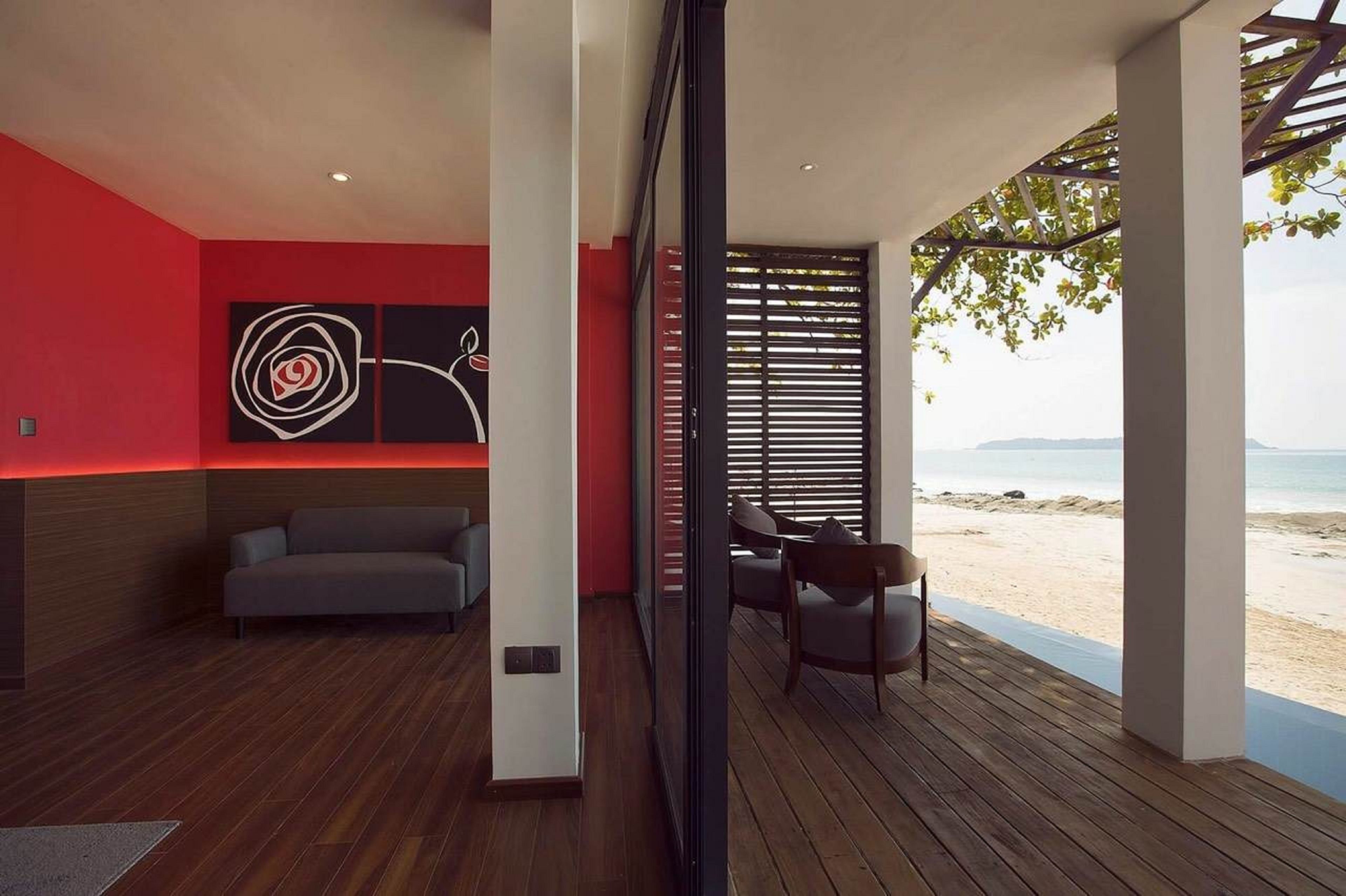 A modern interior with a wooden floor, a red accent wall, a sofa, and a view of the beach.