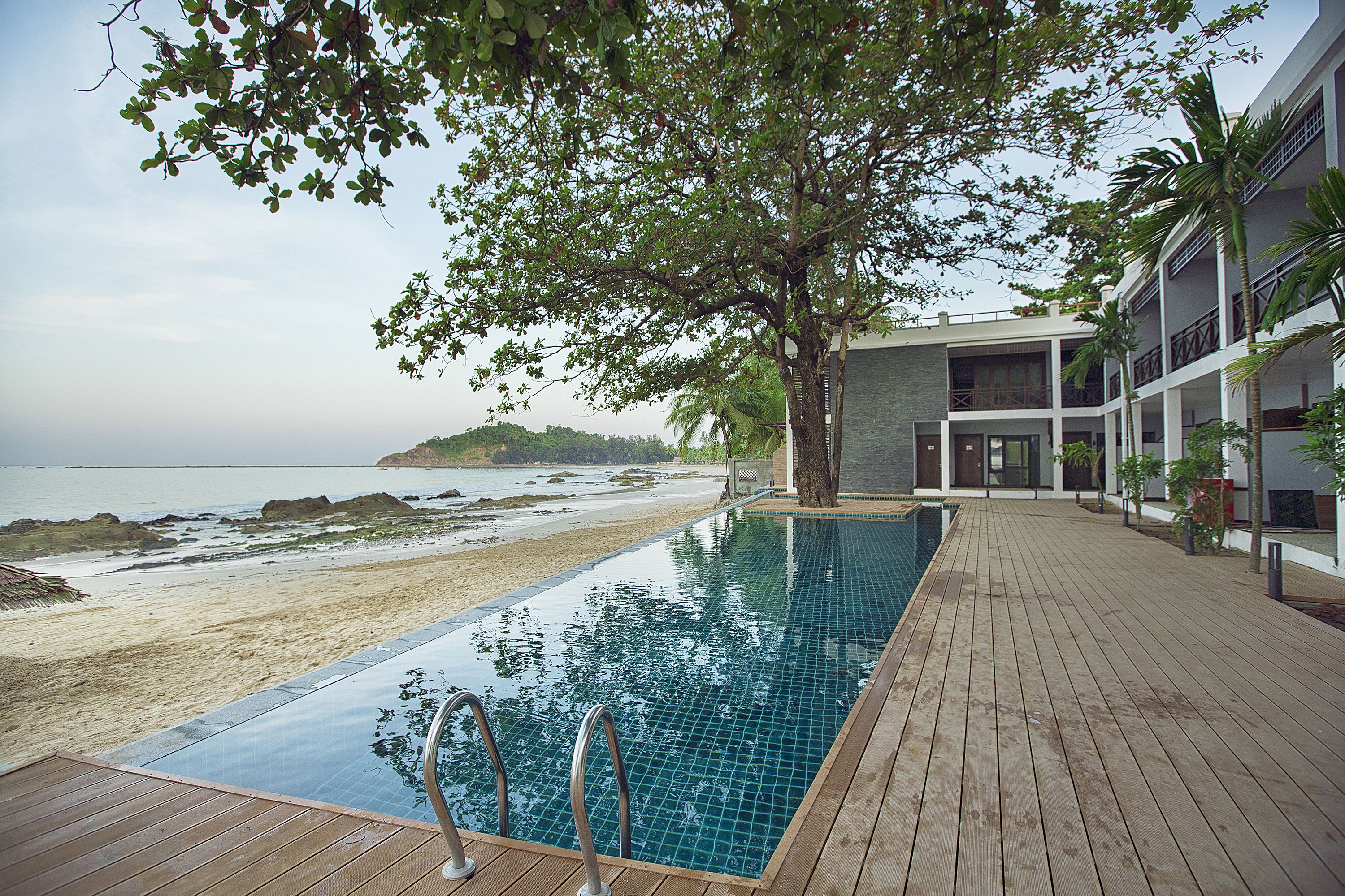 An infinity pool with wooden deck, overlooking a beach with a view of the ocean and a building with balconies.