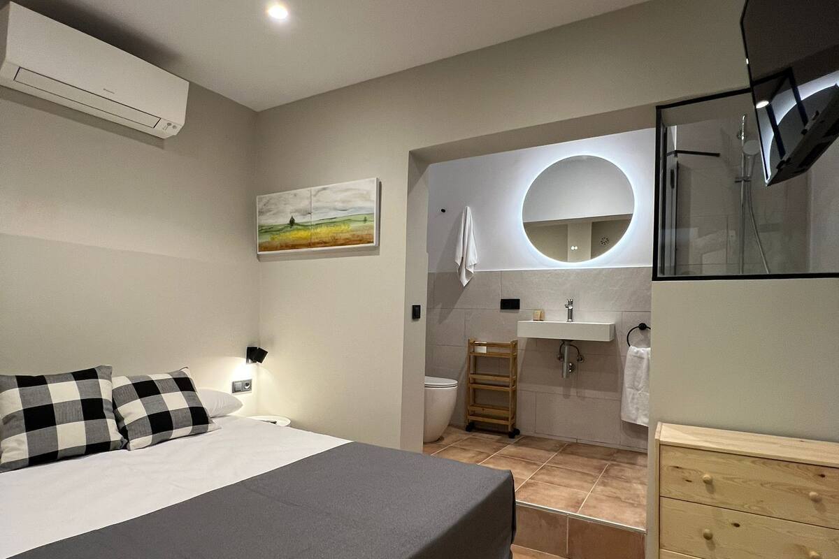 Ein modernes Schlafzimmer mit Bett, Nachttisch, Klimaanlage, einem Gemälde sowie einem Badezimmer mit Waschbecken, Spiegel und Dusche.