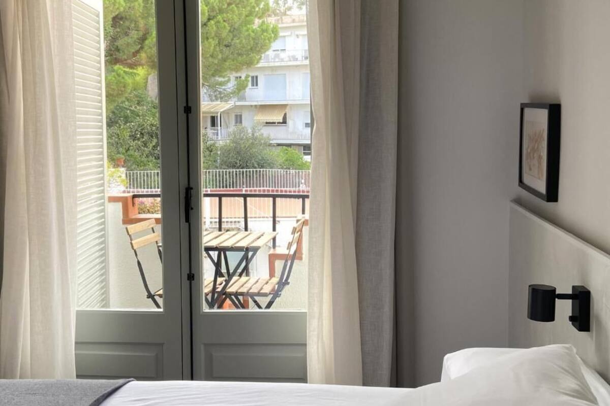 Ein Schlafzimmer mit Bett, Nachttisch, Wandleuchte und Blick auf einen Außenbalkon mit Tisch und Stühlen.