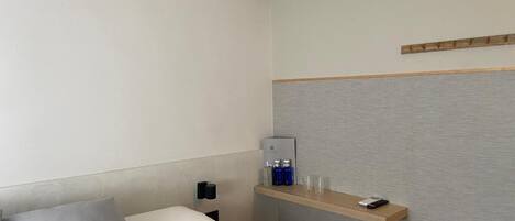 A hotel room with a bed, a bedside table with a lamp, a shelf with toiletries, and an air conditioning unit.