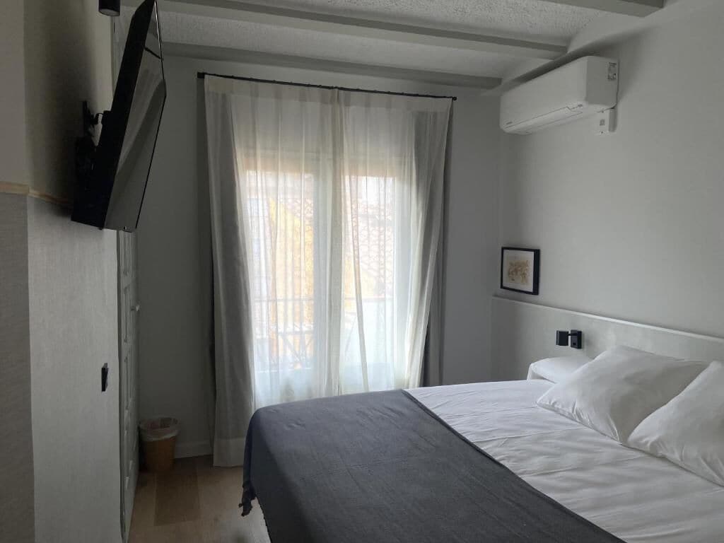 Habitación de hotel con cama, televisor montado en la pared, ventana con cortinas translúcidas y un pequeño cubo de basura.
