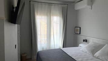 Habitación de hotel con cama, televisor montado en la pared, ventana con cortinas translúcidas y un pequeño cubo de basura.