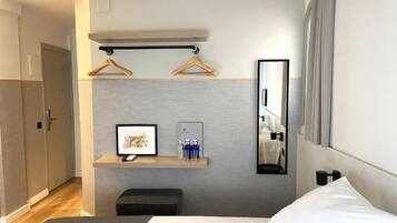 Habitación de hotel con cama, mesita de noche con espejo, perchero y estante pequeño con artículos de aseo.